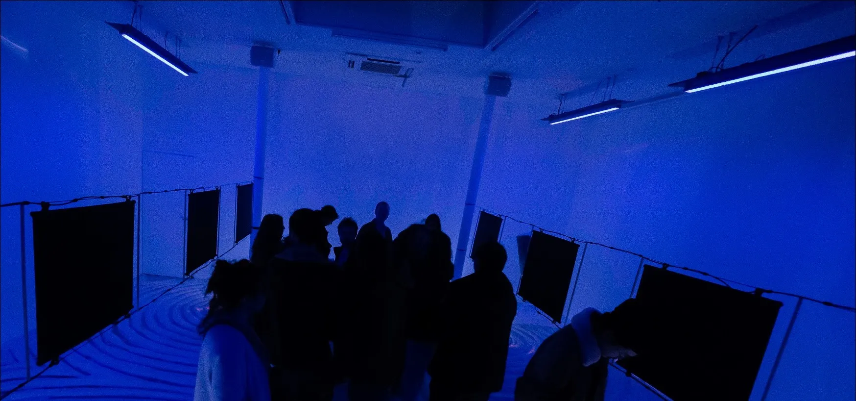 Public dans l'installation Edifice — silhouettes baignées de lumière bleue entre deux rangées de panneaux vibrants suspendus