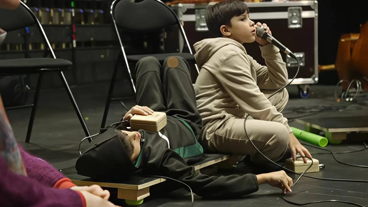 Deux enfants en répétition — l'un allongé avec un Paco posé sur le buste, l'autre chante au micro