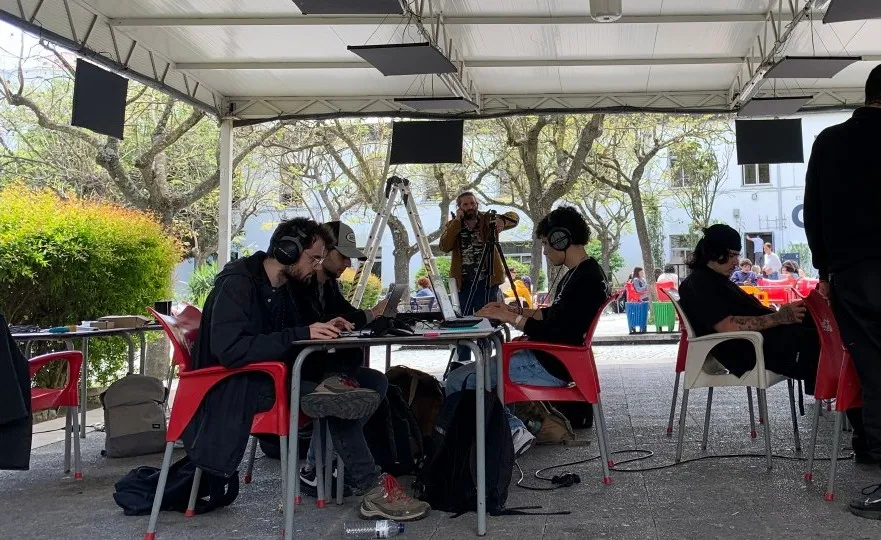 Étudiants au casque autour de tables sous une tonnelle, panneaux vibrants suspendus — workshop AMU à Lisbonne